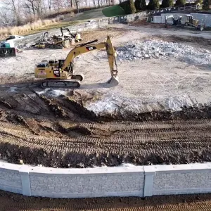 Aerial view of a stripped construction pad inside a curving precast modular block retaining wall, with a tracked excavator at center, a large stockpile of broken concrete and rock rubble beyond it, a skid steer and dozer staged along the far edge, and heavily rutted dark soil across the foreground