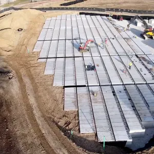 Aerial view of hundreds of numbered pre-placed concrete burial crypts set in long parallel rows across an excavated basin, with workers in high-visibility vests walking the crypt tops, a tracked excavator setting units at the upper right, a wheel loader carrying a crypt along the right edge, and a large soil stockpile at left