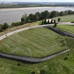 Aerial view of the finished expansion lawn under full irrigation, enclosed by a stone-faced cast retaining wall stepping down the slope, with existing headstone rows on the terraces at right, evergreen plantings along the access road, and the river and a highway bridge in the far distance