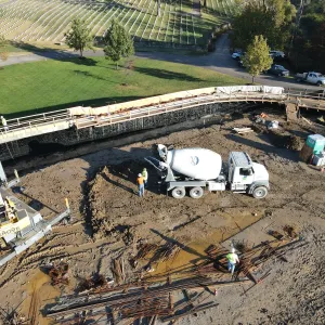 Aerial view of a long curved cast-in-place concrete retaining wall under construction, with exposed vertical reinforcing steel and tarped forms along its length, a concrete mixer truck and two workers on the mud pad below, a rough-terrain crane with boom extended at left, and bundles of reinforcing bar laid out in the foreground