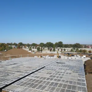 Low aerial view across a completed field of numbered pre-placed crypt lids under a clear blue sky, with an excavator, skid steer, staged individual crypts and a soil stockpile at the far end, and the existing cemetery grounds and treeline on the horizon