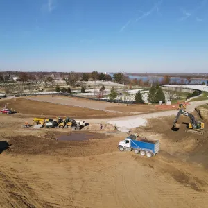Aerial view of mass excavation with a tandem-axle dump truck being loaded at center and a second dump truck at left, a large tracked excavator cutting the bank at right, a lineup of compact excavators, a wheel loader and a water tank trailer on the shelf above, and a muddy water pocket in the base of the cut, with the existing cemetery and river beyond