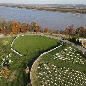 Aerial view of the completed and sodded expansion panel ringed by a white concrete retaining wall and black fence, with newly planted trees in the turf, peak autumn foliage along the bluff, existing headstone sections on both sides, and the Mississippi River filling the upper half of the frame