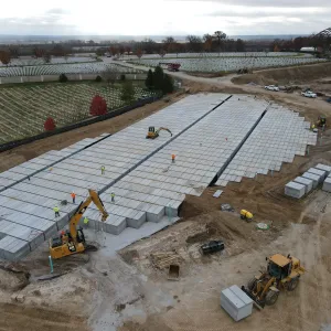 Aerial view of the same pre-placed crypt array under heavy overcast, with workers, an excavator on the deck, a second excavator and a wheel loader moving a crypt at the lower left, staged crypt units and soil stockpiles at right, and an arched steel bridge on the far horizon