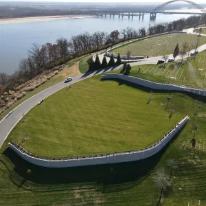 Aerial view of the completed burial expansion panel as a broad striped lawn bounded by a serpentine stone-faced retaining wall with black picket fence and a new loop road, with the existing cemetery terraces at right, the Mississippi River across the top of the frame and an arched steel bridge at the upper right