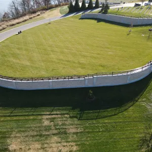 Closer aerial view of the finished expansion lawn and its curving stone-faced retaining wall topped by black picket fence, with young trees in mulch rings, trash receptacles at the road edge, long afternoon shadows across the slope below the wall, and existing headstone rows at the top right
