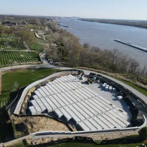 Wide aerial view of the crypt installation area seen from above the bluff, with the partially placed crypt block inside its lined excavation and retaining wall, historic red brick barracks buildings and older headstone sections at left, and a towboat pushing a barge on the Mississippi River at right