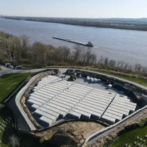 Wide aerial view of the crypt block under installation within the walled and silt-fenced excavation, with staged crypt units and a compact excavator at the top edge, existing headstone rows in the foreground and lower right, and a towboat and barge tow on the river beyond the bare treeline