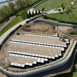 Aerial view of long single rows of pre-placed crypts set on black liner strips across a sand-graded subgrade, with a compact excavator placing units at the right, a pickup truck on the pad at left, the completed retaining wall and silt fence around the perimeter, and existing headstone sections on the terraces above