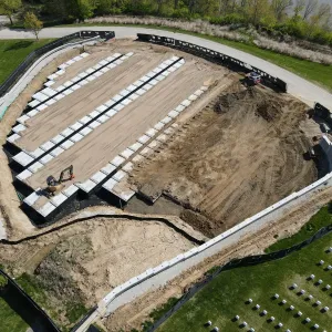 Aerial view of partially installed crypt rows on prepared sand subgrade with a compact excavator working the open end, a service truck at the top edge, large areas of rough-graded and tracked earth to the right, and the new concrete retaining wall curving around the lower perimeter beside existing headstone rows