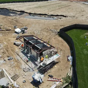 Aerial view of a small restroom building under construction with concrete masonry walls and dark red steel roof framing in place, masons on scaffolding setting stone veneer at the near end, pallets of block and stone and stacked lumber across the surrounding graded site, and a lined bioretention basin with rock outlet protection beyond