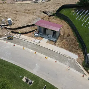 Aerial view of the restroom building with roof underlayment installed and open masonry walls, a walk-behind roller and worker on the new concrete apron, freshly placed concrete curb and aggregate base along the roadway lined with traffic cones, and existing headstone rows behind the fence at right