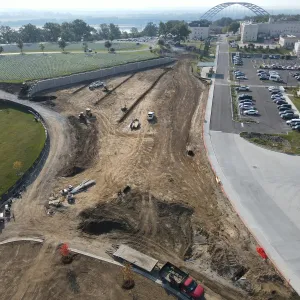 Aerial view of roadway and site grading along a broad earth corridor between the walled expansion area and a completed concrete drive and parking lot serving the adjacent medical center, with a dozer, service trucks and stacked pipe on the graded ground, and the cemetery terraces and an arched bridge in the distance