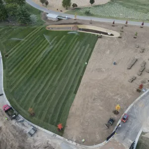 Aerial view of freshly sodded and irrigated lawn with sprinklers running across the striped turf, pallets of sod rolls staged on the bare graded soil at right, a newly built bioretention basin with mulch and rock at the top, young trees in mulch rings, and a crew and equipment along the perimeter roads