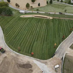 Aerial view of the fully sodded expansion area with mowing stripes across the turf, a completed bioretention basin with riprap outlet at the center, new trees along the roadways, a dump truck and equipment on the drive at left, and existing headstone sections and a committal shelter beyond