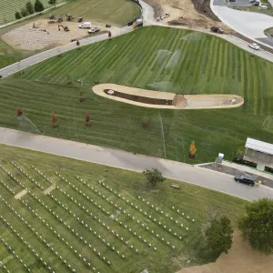 Aerial view of the finished turf areas under irrigation with sprinklers arcing over the lawn, a lined bioretention basin with riprap and turf reinforcement matting at the center, a new open committal shelter with white metal roof beside a coned-off drive at right, and dense existing headstone rows filling the foreground