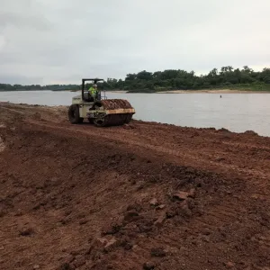 Pad-foot roller compacting red clay fill along the embankment crest beside the river