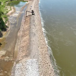Aerial view of a long-reach excavator and haul truck on the crest, with a patch of newly placed light riprap at the toe