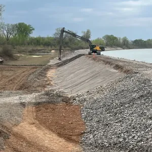 Excavator at the top of a shaped embankment section covered with geotextile fabric, with stone stockpiled alongside