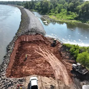 Aerial view of an exposed section of bare fill between armored reaches, with a dozer, a smooth-drum roller, workers and a pickup