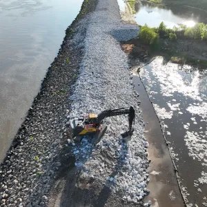 Excavator placing riprap along the crest and slope in low sun, with silt fence along the landside mudflat