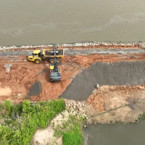 Articulated dump truck and excavator working over a gray stone pad, with a worker holding a survey rod