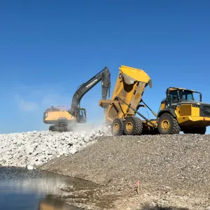 Excavator and articulated dump truck dumping and spreading light-colored riprap on the crest against a clear sky