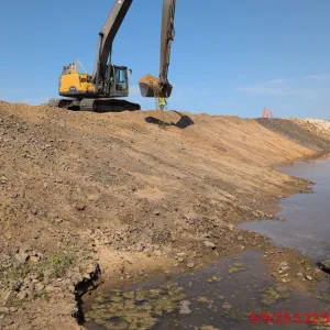Excavator dumping a bucket of soil onto a graded slope beside standing water, with erosion-control tubes staked along the far bank
