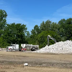 Tractor-trailer with a belly-dump trailer beside a large stone stockpile being worked by an excavator in the staging area